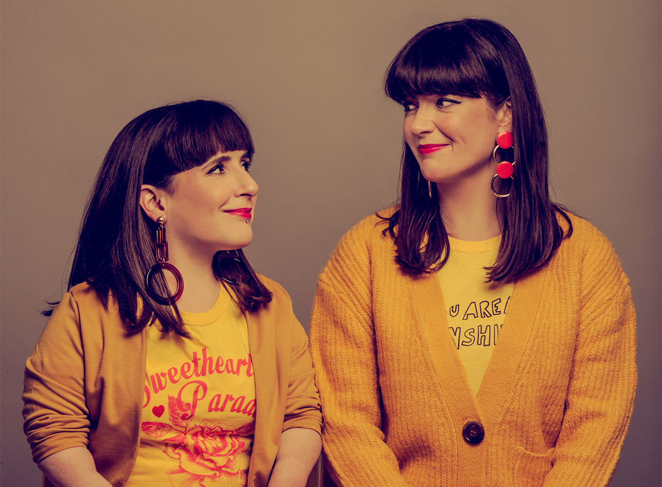 Two women looking at eachother smiling with similar yellow jackets, hair and earrings.